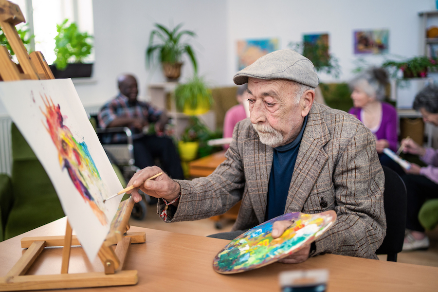 An elderly man is creating a colorful picture, using paintbrush and palette for a recreational activity in an assisted living retirement home