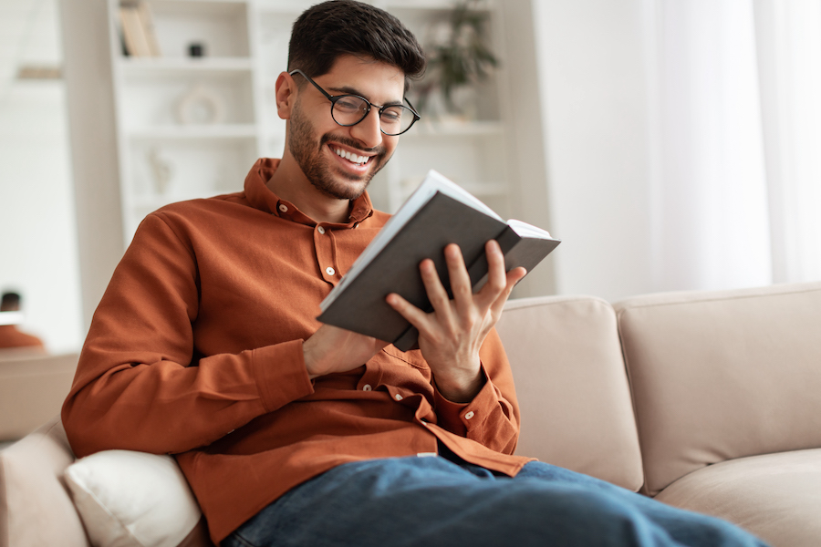 Leisure and Relaxation Concept. Portrait of a cheerful smiling young Middle Eastern man taking a break from caregiving wearing eyeglasses holding paper book, sitting on couch at home in living room, reading literature, enjoying story.